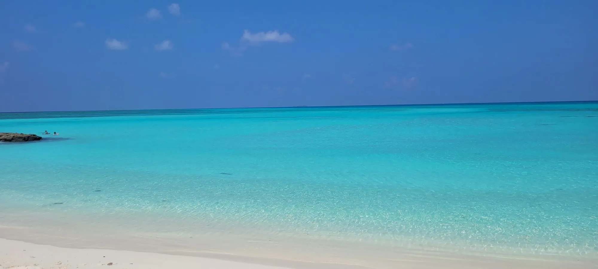 Heller Sandstrand am Bikini Beach von Fulidhoo, davor glasklares türkises Flachwasser bis zum Horizont, links ein kleiner Felsen.