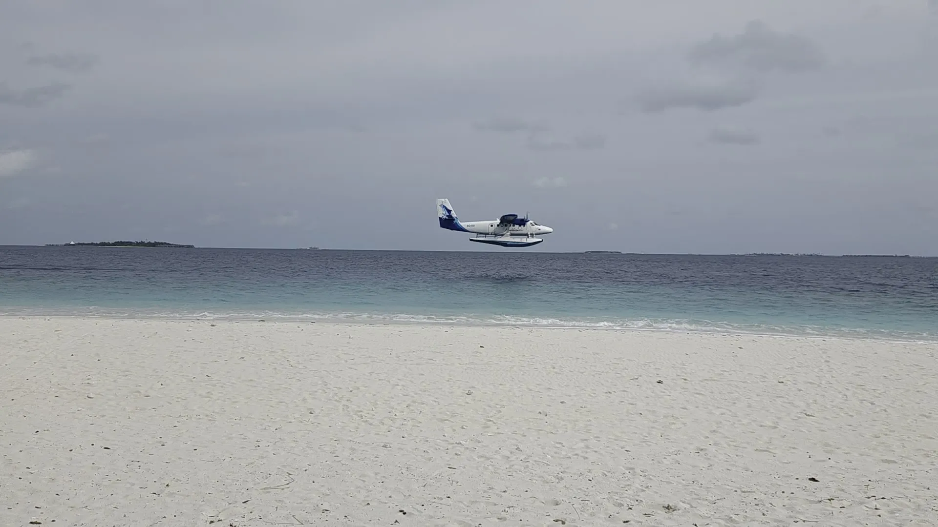 Ein weiß-blaues Wasserflugzeug hebt vor dem breiten weißen Sandstrand von Kamadhoo vom Meer ab, dahinter das offene Wasser und eine bewaldete Insel am Horizont.