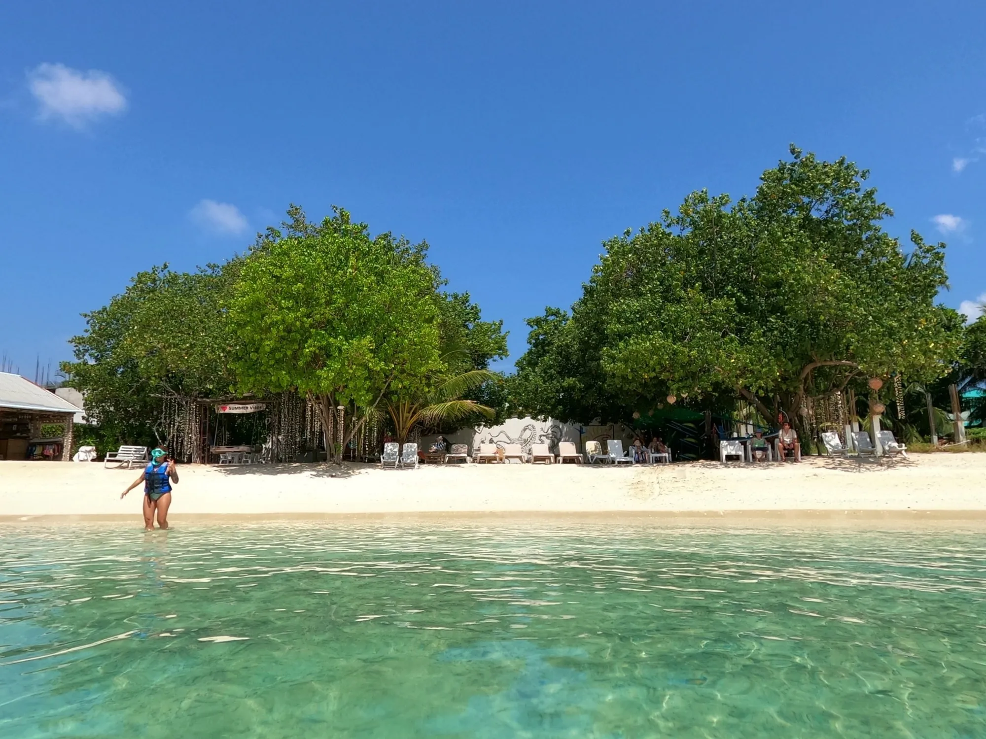 Blick vom Wasser auf den Bikini-Strand von Rasdhoo: heller Sand, große Schattenbäume und Liegestühle, im Flachwasser steht eine Schnorchlerin mit Maske und Schwimmweste.