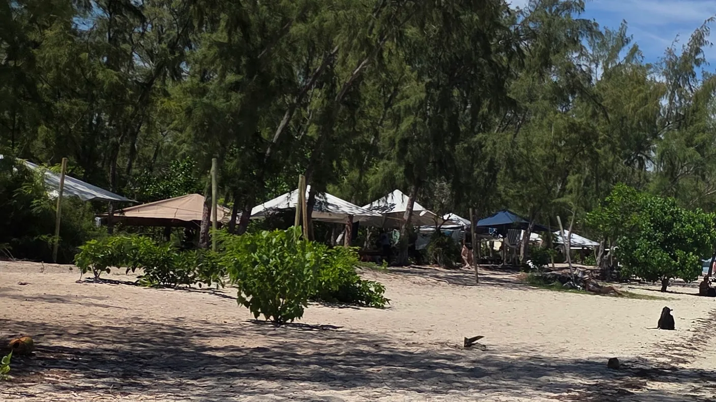 Heller Strand der Île aux Bénitiers, dahinter Bäume und kleine Stände im Schatten